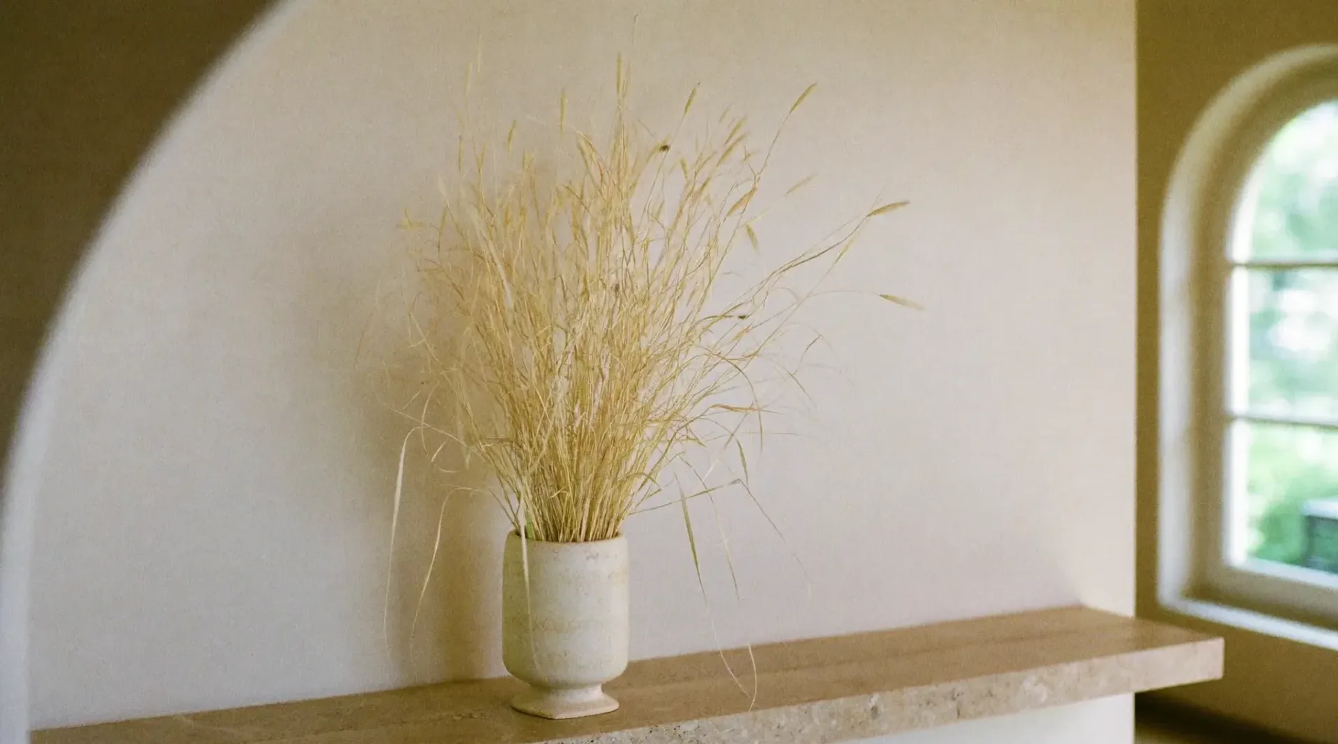 Haz de tallos de pasto seco en un vaso de travertino sobre una mesada de piedra, con la sombra de una ventana en arco en la pared