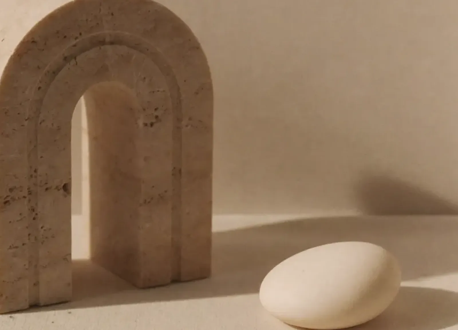 Pequeno arco de travertino de pé sobre linho cor de areia, com uma pedra lisa ao lado, contra uma parede de reboco marfim