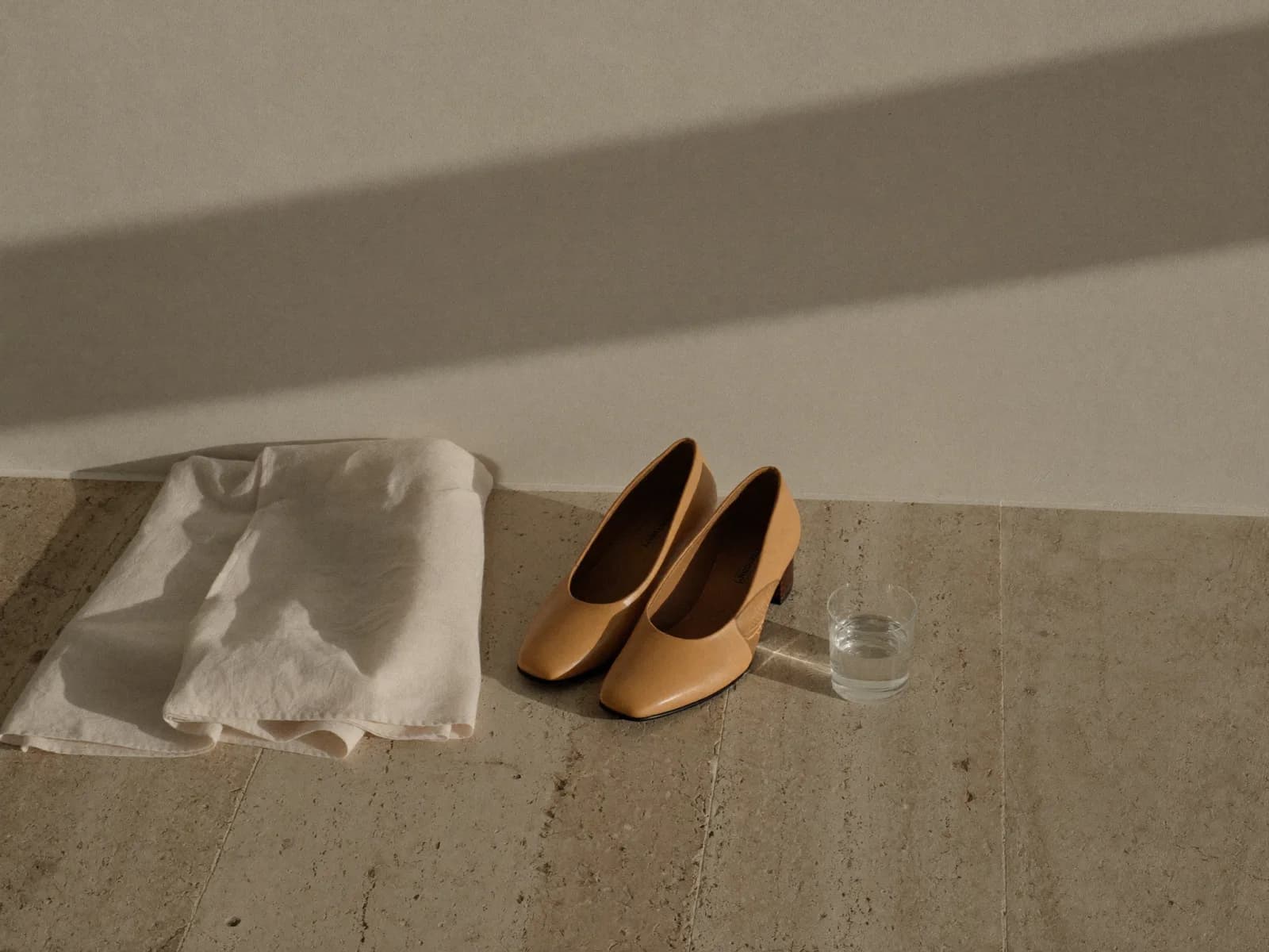 Par de zapatos bajos color arena sobre piso de travertino, junto a un paño de lino doblado y un vaso de agua, con la sombra de una ventana en la pared