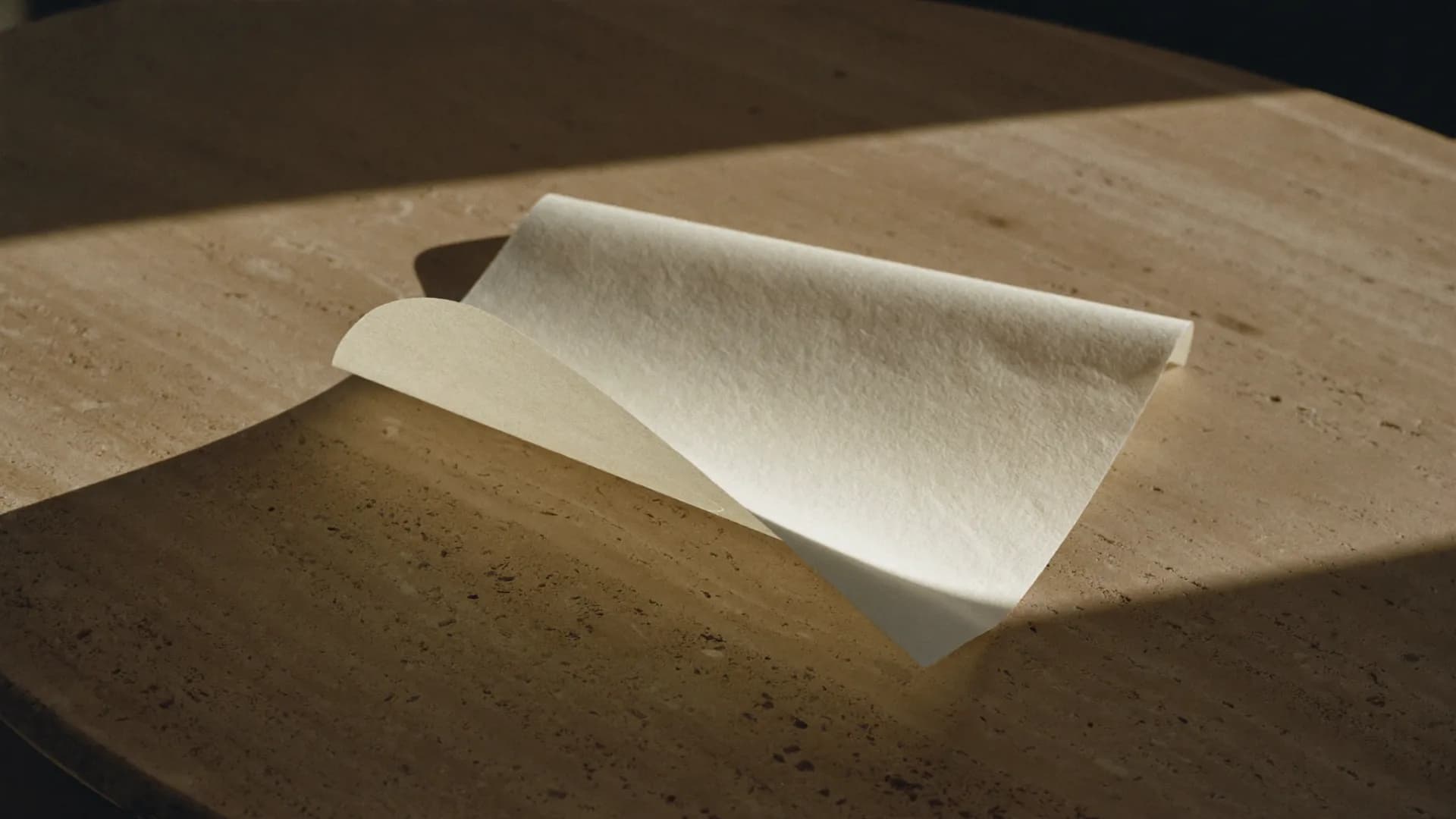 Hoja de papel artesanal color marfil, levemente curvada, sobre una mesada de travertino con luz cálida de ventana